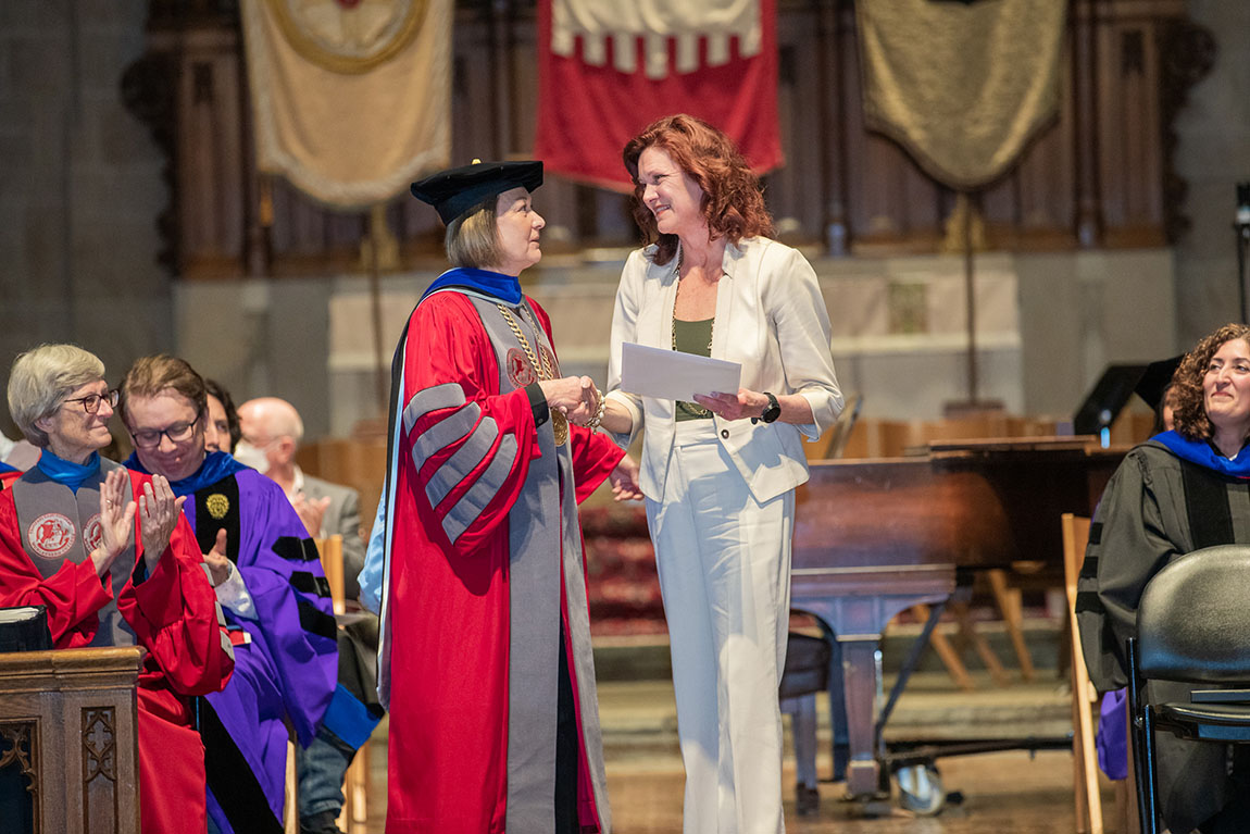 A woman in academic robes and a hat shakes hands with a woman with long hair wearing a white pantsuit